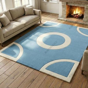 May include: A rectangular blue area rug with a large white circle in the center and white geometric shapes at the corners. The rug is placed on a hardwood floor in a living room setting.