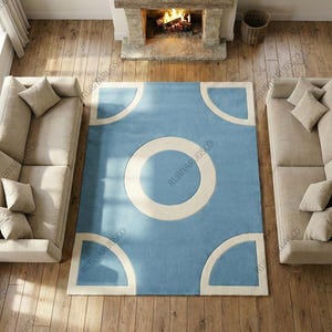 May include: A rectangular blue rug with a large off-white circle in the center and four quarter-circle shapes in the corners. The rug is placed on a wooden floor, with two beige sofas on either side. A fireplace is visible in the background.