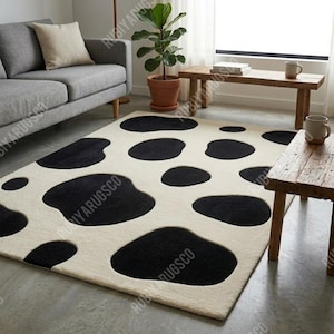 May include: A cream-coloured rectangular rug featuring large, irregular black spots. The rug is positioned on a light-coloured floor in a living room, with a sofa and wooden furniture visible in the background. The rug's design resembles a cowhide pattern.