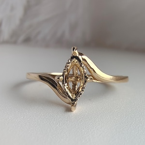 May include: A gold-colored ring with a unique design. The ring features a central, diamond-shaped element with a cross-like pattern, flanked by curved bands. The ring is set against a white surface with a soft, feathery background.