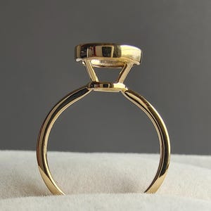May include: A gold ring featuring a smooth, thin band and a raised setting that holds a single gemstone. The metal has a polished, reflective finish, and the design includes a basket-style gallery that supports the stone from underneath.