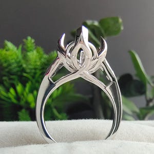 May include: A polished silver-toned ring featuring a complex, multi-pronged setting that rises to a central point. The band is smooth and tapers toward the base, while the upper section displays an intricate, open-work design with curved metal elements.