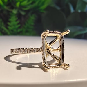 May include: A gold ring setting featuring a rectangular prong head designed to hold a center stone. The band is adorned with a row of small, round, clear pavé-set stones along the shank, reflecting light with a polished metallic finish.