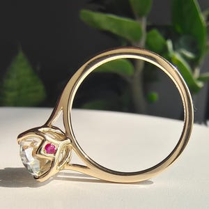 May include: A gold engagement ring with a large, clear gemstone and a small, red gemstone accent. The ring is set on a white surface, with green foliage in the background. The ring's band is a simple, elegant design.