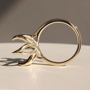 May include: A gold-colored ring with a unique floral design. The ring features a circular band with a stylized three-petal flower design on one side. The ring is displayed on a white surface with a neutral background, showcasing its intricate details.
