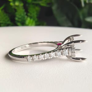 May include: A silver-toned ring featuring a row of small, clear, round-cut stones along the band. The setting includes a prominent prong structure designed to hold a center stone, accented by a single, small, round, vibrant red gemstone.