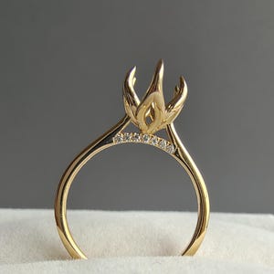 May include: A gold ring featuring a high, tulip-shaped prong setting designed to hold a center stone. The band is accented with a row of small, clear pavé-set diamonds along the upper curve, leading up to the intricate, petal-like metal prongs.