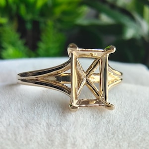 May include: A polished gold ring setting featuring a rectangular four-prong head designed to hold a gemstone. The band has a split shank design that tapers toward the center, showcasing a smooth, reflective metallic finish throughout the structure.