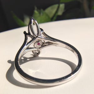 May include: A silver-toned ring featuring an intricate openwork design on the band. The interior of the band is clearly stamped with the text S925. A small, circular, deep pink gemstone is set within the decorative metalwork of the ring setting.
