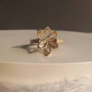 May include: A gold-colored ring with a floral design. The ring features an openwork flower with six petals. The band has an engraved pattern. The ring is displayed on a white surface with a soft, blurred background.