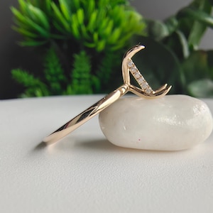 May include: A gold ring featuring a crescent moon-shaped setting. The curved metal frame is accented with a row of small, clear, round-cut stones embedded along the inner edge of the crescent, creating a delicate and refined appearance.