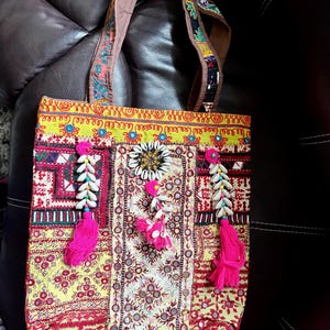 Può includere: Una colorata borsa tote con un design patchwork. La borsa presenta un motivo giallo, rosso e bianco con nappe rosa e dettagli di conchiglie. I manici sono marroni con dettagli ricamati. Al centro c'è un fiore decorativo.