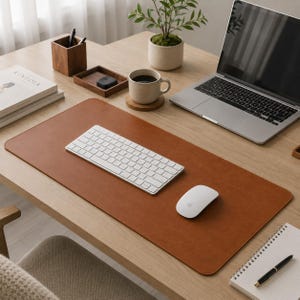 Pode incluir: Um tapete de secretária retangular de cor castanha com uma superfície lisa, apresentando um teclado sem fios branco e um rato de computador sem fios branco por cima. O tapete está posicionado numa secretária de madeira com um portátil.