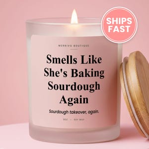 May include: A frosted glass candle with a light pink label featuring the text Merrivo Boutique, Smells Like She's Baking Sourdough Again, Sourdough takeover, again, and 8oz Soy Wax. The candle has a lit wick and a separate light brown bamboo lid.