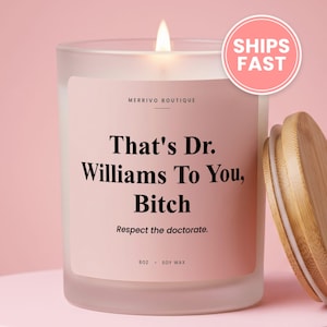 May include: A frosted glass candle in a jar with a light pink label featuring the black text: Merrivo Boutique, That's Dr. Williams To You, Bitch, Respect the doctorate, 8oz, soy wax. The candle has a lit wick and a separate light wood lid.