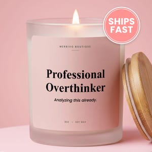 May include: A frosted glass candle with a light pink label featuring the text Merrivo Boutique, Professional Overthinker, Analyzing this already, and 8oz Soy Wax. The candle has a lit wick and is accompanied by a separate light wood lid.
