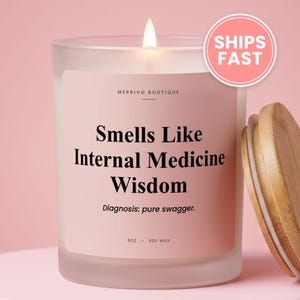 May include: A frosted glass candle in a jar with a light pink label featuring the text Merrivo Boutique, Smells Like Internal Medicine Wisdom, Diagnosis: pure swagger, 8oz, and soy wax. A lit wick and a detached wooden lid are visible.