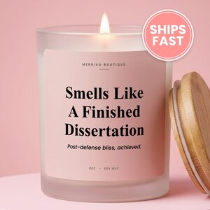 May include: A frosted glass candle with a light pink label featuring the text Merrivo Boutique, Smells Like A Finished Dissertation, Post-defense bliss, achieved, and 8oz Soy Wax. The candle has a lit wick and a separate light wood lid.
