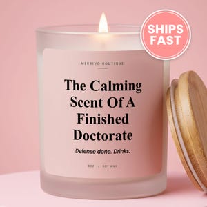 May include: A frosted glass candle with a light pink label featuring the text MERRIVO BOUTIQUE, The Calming Scent Of A Finished Doctorate, Defense done. Drinks., 8oz, and SOY WAX. The candle has a lit wick and a separate light brown bamboo lid.