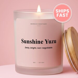 May include: A frosted glass candle with a light pink label featuring the text Merrivo Boutique, Sunshine Yuzu, Zesty, bright, non-negotiable, 8oz, and soy wax. A lit wick burns at the top, and a round bamboo lid sits to the right of the candle.