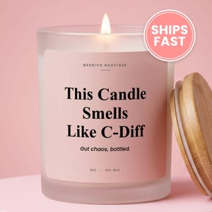 May include: A frosted glass candle with a light pink label featuring the text Merrivo Boutique, This Candle Smells Like C-Diff, Gut chaos, bottled, 8oz, and soy wax. The candle has a lit wick and is accompanied by a round bamboo lid.