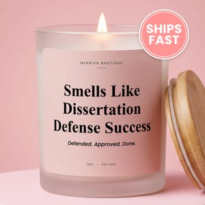 May include: A frosted glass candle with a light pink label featuring the text Merrivo Boutique, Smells Like Dissertation Defense Success, Defended. Approved. Done. 8oz. Soy Wax. The candle has a lit wick and a separate light brown wooden lid.