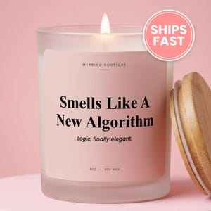 May include: A frosted glass candle in a jar with a light pink label featuring the text Merrivo Boutique, Smells Like A New Algorithm, Logic, finally elegant, 8oz, and soy wax. A wooden lid sits to the right of the candle with a lit wick.