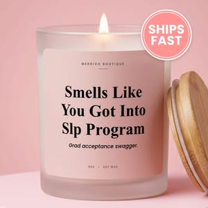 May include: A frosted glass candle in a jar with a light pink label featuring the text "Smells Like You Got Into Slp Program" and "Grad acceptance swagger." The candle has a lit wick and is accompanied by a separate light-colored wooden lid.