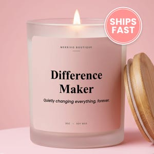 May include: A frosted glass candle with a light pink label featuring the text Merrivo Boutique, Difference Maker, and Quietly changing everything, forever. The label also states 8oz and soy wax. A bamboo lid sits beside the candle.