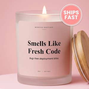 May include: A frosted glass candle with a light pink label featuring the text Merrivo Boutique, Smells Like Fresh Code, Bug-free deployment bliss, and 8oz Soy Wax. The candle has a lit wick and is accompanied by a round bamboo lid.