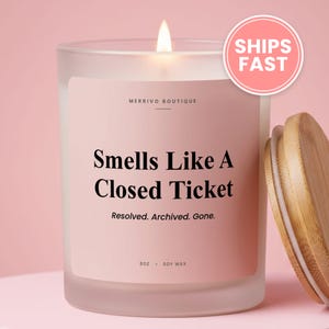 May include: A frosted glass candle with a light pink label featuring the text Merrivo Boutique, Smells Like A Closed Ticket, Resolved. Archived. Gone. 8oz. Soy Wax. The candle has a lit wick and is accompanied by a separate light brown wooden lid.