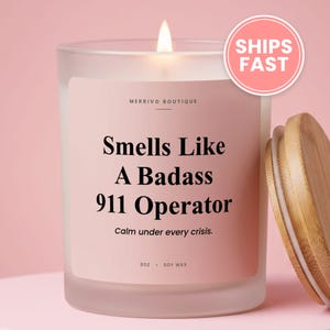 May include: A frosted glass candle with a light pink label featuring the text Merrivo Boutique, Smells Like A Badass 911 Operator, Calm under every crisis, 8oz, Soy Wax. The candle has a lit wick and a separate light brown wooden lid.
