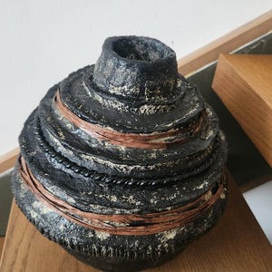Può includere: Un vaso decorativo fatto a mano, di colore scuro, con una superficie strutturata. Il vaso presenta un design a strati con fasce di accenti marroni e neri a forma di corda. Il vaso è esposto su una superficie di legno.