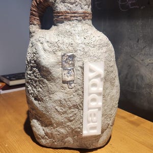 Può includere: Una brocca grigia strutturata con un manico intrecciato e un'etichetta rettangolare bianca con la scritta "happy". La brocca ha un piccolo ornamento metallico quadrato. La brocca è appoggiata su una superficie di legno.