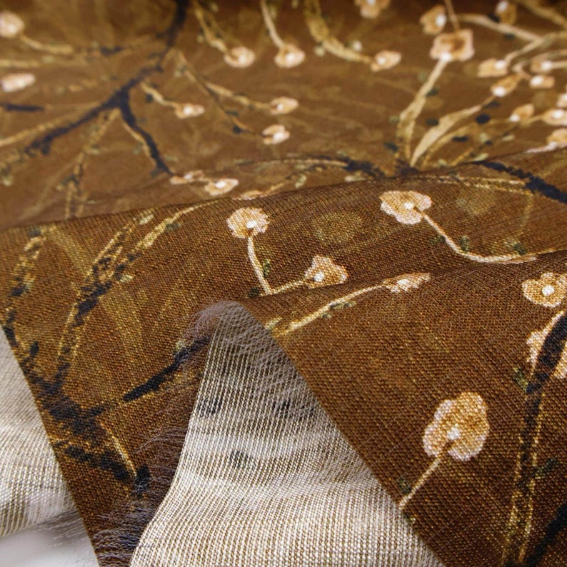 以下が含まれることがあります： 茶色の生地のクローズアップ。白い花と黄色の花、細い茎と濃い茶色の枝が描かれたデザイン。生地は質感があり、折りたたまれた部分には明るい色の裏面が見えます。