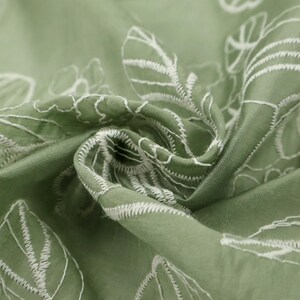 May include: A sage green fabric featuring a white embroidered botanical pattern of leaves and flowers. The material is gathered in the center, creating folds and shadows that highlight the texture of the stitching and the smooth fabric surface.