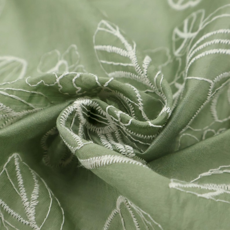 May include: A sage green fabric featuring a white embroidered botanical pattern of leaves and flowers. The material is gathered in the center, creating folds and shadows that highlight the texture of the stitching and the smooth fabric surface.