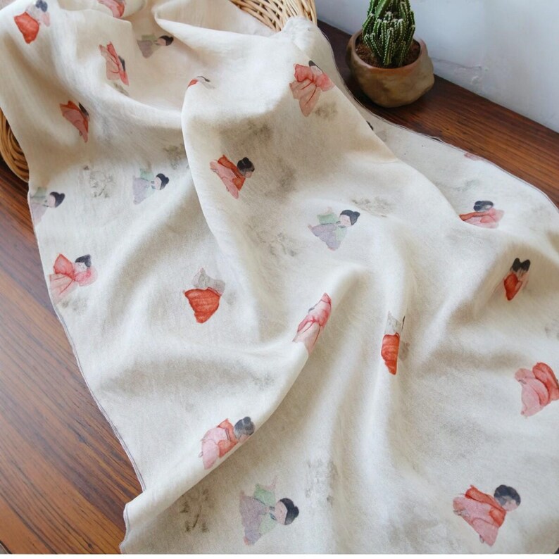 Peut inclure: &Eacute;charpe cr&egrave;me avec un motif r&eacute;p&eacute;t&eacute; de personnages &agrave; l'aquarelle rouges, roses et gris. L'&eacute;charpe est drap&eacute;e sur un panier en osier et une surface en bois, avec un petit cactus en pot en arri&egrave;re-plan.