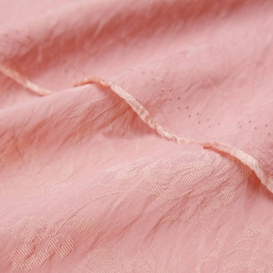 May include: A light pink fabric featuring a subtle, tonal floral jacquard pattern woven into the material. The textile has a crinkled, textured surface with a delicate, folded edge that displays a fine row of small, evenly spaced perforations.