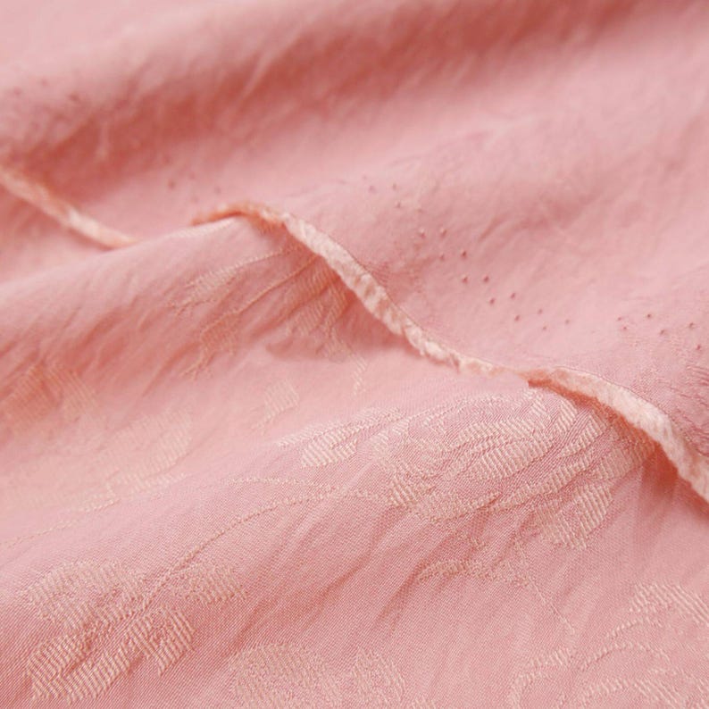 May include: A light pink fabric featuring a subtle, tonal floral jacquard pattern woven into the material. The textile has a crinkled, textured surface with a delicate, folded edge that displays a fine row of small, evenly spaced perforations.