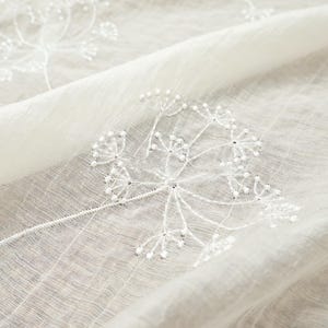 May include: Close-up of a sheer, off-white fabric with embroidered white floral designs. The delicate embroidery features detailed seed head patterns. The fabric has a subtle grid pattern and is gently folded, creating soft shadows.