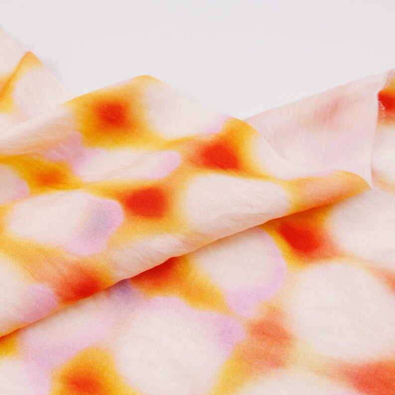May include: Close-up of a folded fabric with a pattern of orange, yellow, and pink spots on a white background. The fabric appears to be lightweight and flowing, with visible folds and creases.