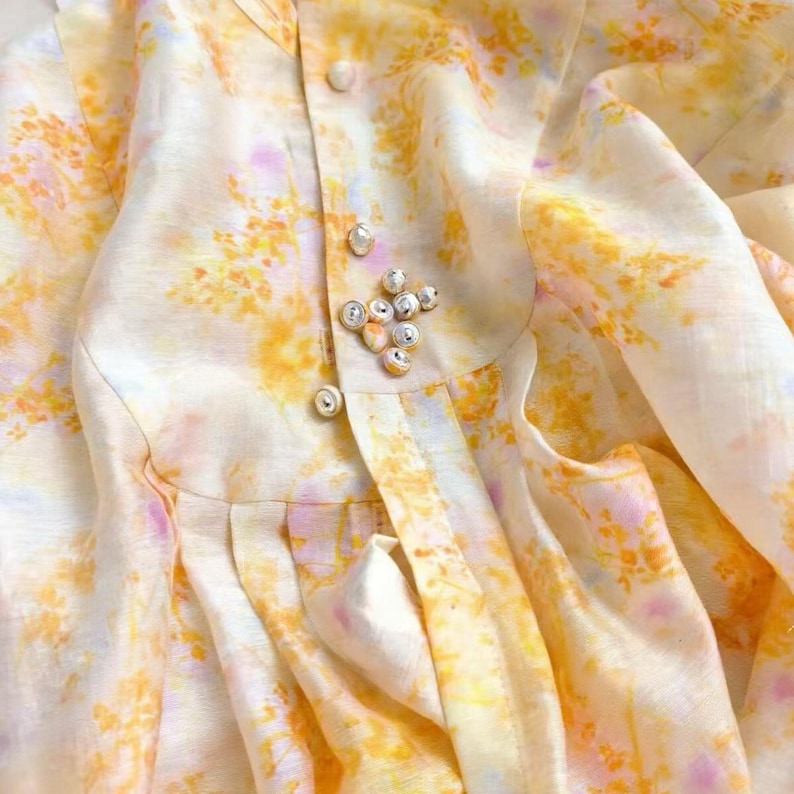 May include: Close-up of a cream-colored garment with a floral pattern in yellow, orange, and pink. The garment features a button-up closure with pearl-like buttons and a decorative brooch.