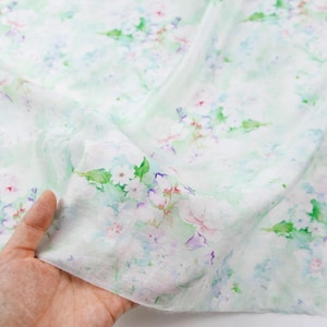 May include: A piece of fabric with a delicate floral pattern. The design features watercolor-style flowers in shades of white, pink, purple, and green, set against a soft, pale green background. The fabric is held in a hand, showing its texture and drape.