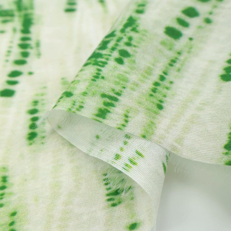 Op de afbeelding: Een stuk gebroken wit textiel met een verticaal tie-dye patroon in tinten levendig groen. De stof heeft een semi-transparante, lichtgewicht uitstraling met zichtbare geweven vezels en zachte, gevouwen randen die het kleurverloop benadrukken.
