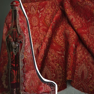 May include: A vibrant red fabric featuring an intricate gold and dark red paisley pattern. The textile displays a rich, textured weave with metallic gold threads woven throughout the design, creating a complex, repeating floral and teardrop motif.