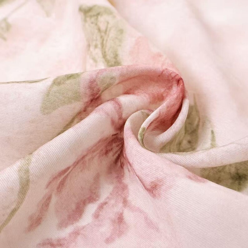 May include: Close-up of a light pink fabric with a floral pattern. The design features large, watercolor-style flowers in shades of pink and red, with green leaves. The fabric is soft and appears to be draped.