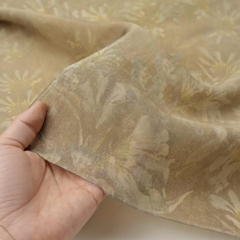 Puede incluir: Una tela ligera y transparente en una paleta de colores beige y bronceado apagado. El tejido presenta un patr&oacute;n floral sutil y repetitivo con formas de p&eacute;talos suaves y superpuestas en tonos crema m&aacute;s claros, creando un aspecto delicado.