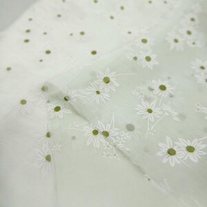 May include: A sheer, light green fabric featuring a repeating pattern of white daisy flowers with yellow-green centers. The material has a translucent, lightweight quality with a soft texture, showcasing the floral print across the surface.
