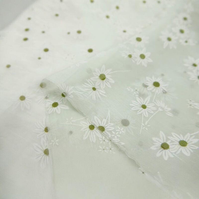 May include: A sheer, light green fabric featuring a repeating pattern of white daisy flowers with yellow-green centers. The material has a translucent, lightweight quality with a soft texture, showcasing the floral print across the surface.